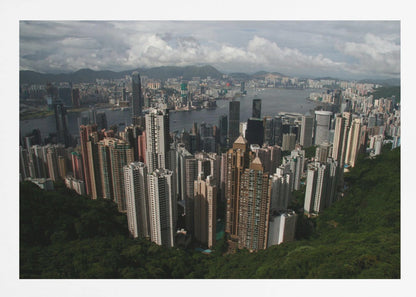 A high-angle photograph of the Hong Kong cityscape, viewed from a lush green peak. Numerous skyscrapers fill the frame, overlooking a bustling harbor with mountains in the distant background under a cloudy sky. The entire image is presented within a silver, brushed-metal frame. Artwork