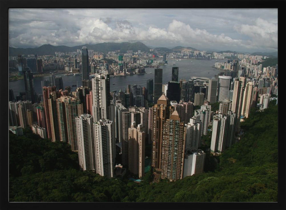 A high-angle photograph of the Hong Kong cityscape, viewed from a lush green peak. Numerous skyscrapers fill the frame, overlooking a bustling harbor with mountains in the distant background under a cloudy sky. The entire image is presented within a silver, brushed-metal frame. Artwork