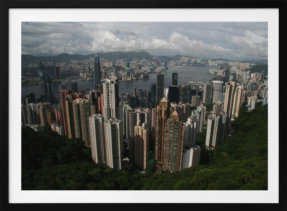 A high-angle photograph of the Hong Kong cityscape, viewed from a lush green peak. Numerous skyscrapers fill the frame, overlooking a bustling harbor with mountains in the distant background under a cloudy sky. The entire image is presented within a silver, brushed-metal frame. Artwork