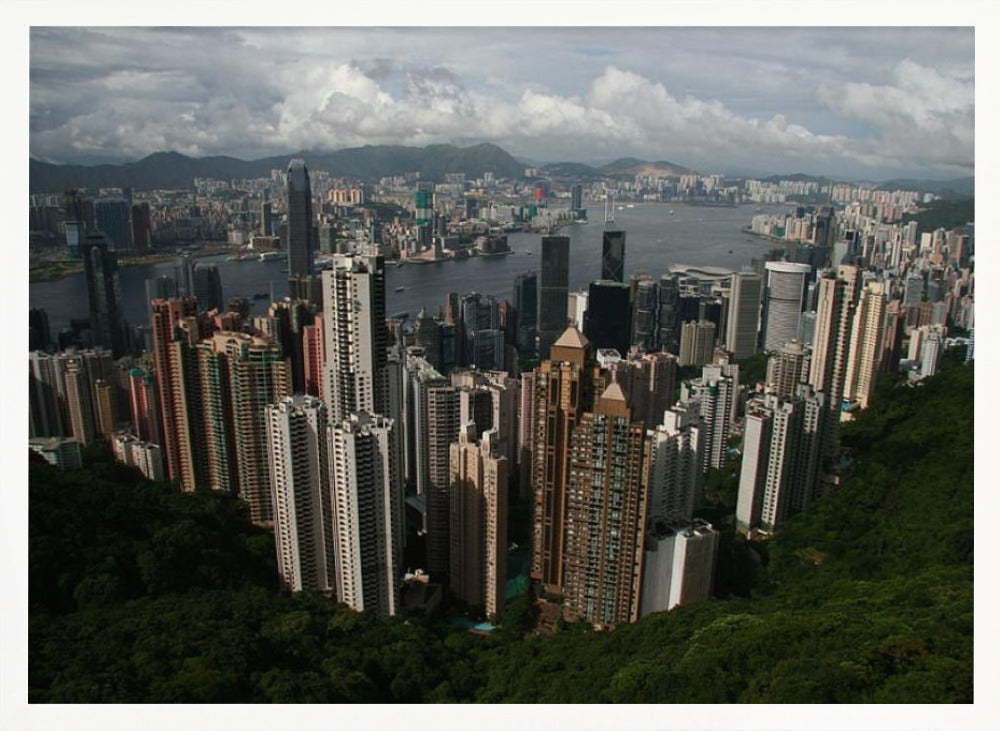 A high-angle photograph of the Hong Kong cityscape, viewed from a lush green peak. Numerous skyscrapers fill the frame, overlooking a bustling harbor with mountains in the distant background under a cloudy sky. The entire image is presented within a silver, brushed-metal frame. Artwork