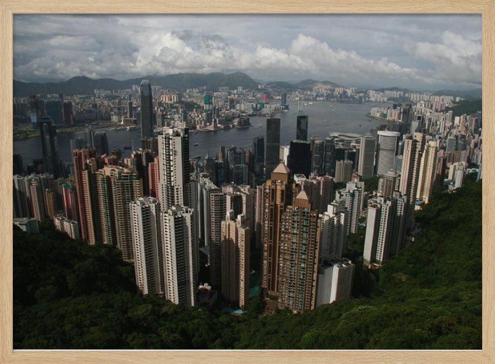 A high-angle photograph of the Hong Kong cityscape, viewed from a lush green peak. Numerous skyscrapers fill the frame, overlooking a bustling harbor with mountains in the distant background under a cloudy sky. The entire image is presented within a silver, brushed-metal frame. Artwork