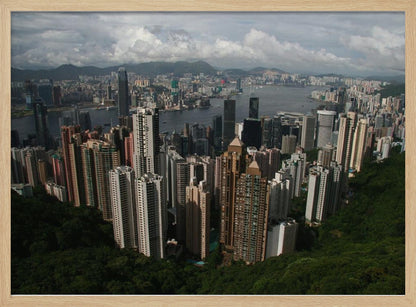 A high-angle photograph of the Hong Kong cityscape, viewed from a lush green peak. Numerous skyscrapers fill the frame, overlooking a bustling harbor with mountains in the distant background under a cloudy sky. The entire image is presented within a silver, brushed-metal frame. Artwork