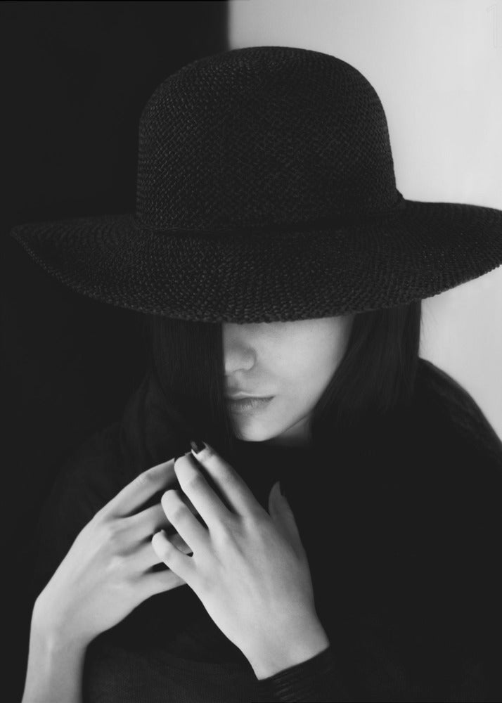 A mysterious black and white portrait of a woman wearing a large, black, wide-brimmed hat that casts a deep shadow over her eyes. Her hands are clasped at her chest, and the background is split vertically into black and white sections. Artwork