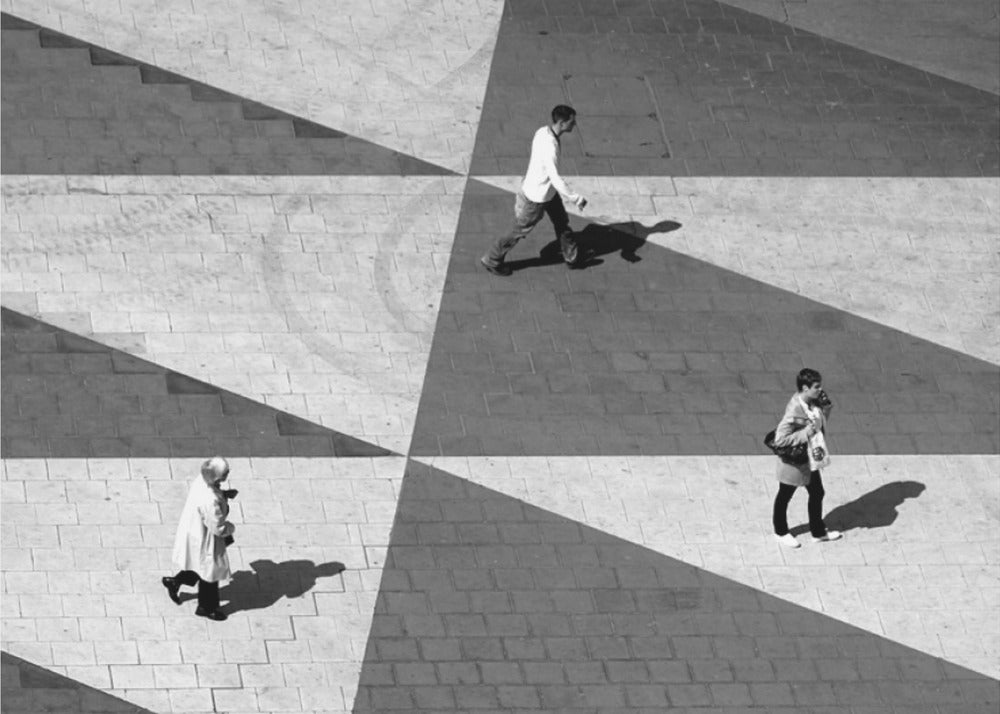 A high-angle, black and white photograph capturing three people walking across a paved plaza, their paths intersecting with a dramatic geometric pattern of light and shadow. The strong sunlight creates large, sharp-edged triangles of shade, isolating each figure in the composition, which is enclosed in a silver frame. Decor