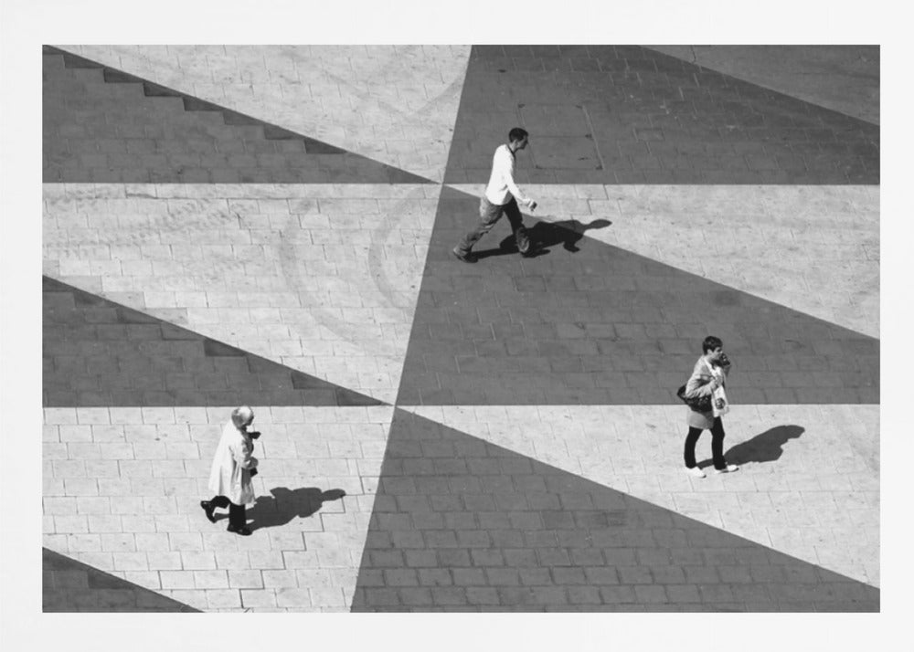 A high-angle, black and white photograph capturing three people walking across a paved plaza, their paths intersecting with a dramatic geometric pattern of light and shadow. The strong sunlight creates large, sharp-edged triangles of shade, isolating each figure in the composition, which is enclosed in a silver frame. Decor