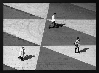 A high-angle, black and white photograph capturing three people walking across a paved plaza, their paths intersecting with a dramatic geometric pattern of light and shadow. The strong sunlight creates large, sharp-edged triangles of shade, isolating each figure in the composition, which is enclosed in a silver frame. Decor