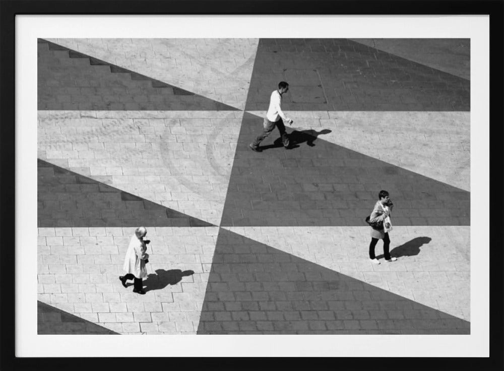A high-angle, black and white photograph capturing three people walking across a paved plaza, their paths intersecting with a dramatic geometric pattern of light and shadow. The strong sunlight creates large, sharp-edged triangles of shade, isolating each figure in the composition, which is enclosed in a silver frame. Decor
