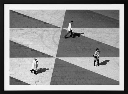 A high-angle, black and white photograph capturing three people walking across a paved plaza, their paths intersecting with a dramatic geometric pattern of light and shadow. The strong sunlight creates large, sharp-edged triangles of shade, isolating each figure in the composition, which is enclosed in a silver frame. Decor