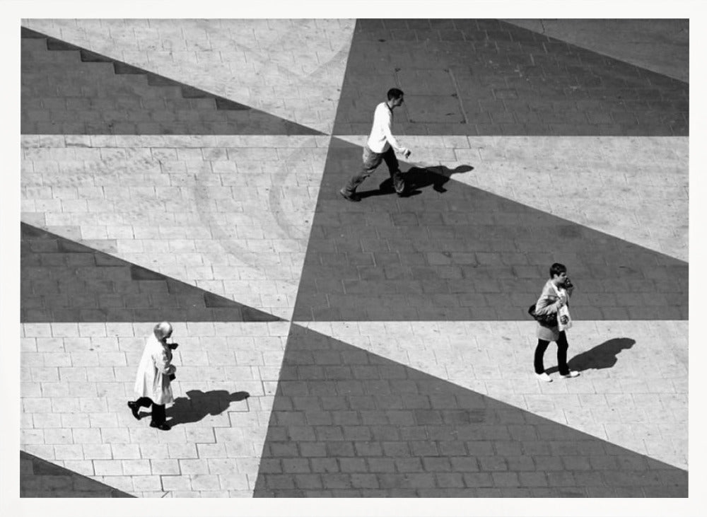 A high-angle, black and white photograph capturing three people walking across a paved plaza, their paths intersecting with a dramatic geometric pattern of light and shadow. The strong sunlight creates large, sharp-edged triangles of shade, isolating each figure in the composition, which is enclosed in a silver frame. Decor