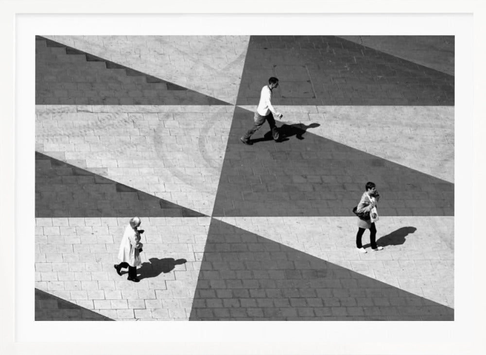 A high-angle, black and white photograph capturing three people walking across a paved plaza, their paths intersecting with a dramatic geometric pattern of light and shadow. The strong sunlight creates large, sharp-edged triangles of shade, isolating each figure in the composition, which is enclosed in a silver frame. Decor