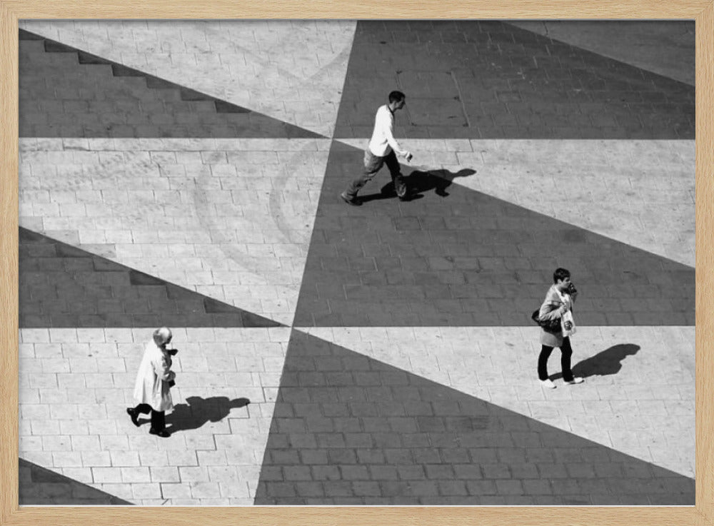 A high-angle, black and white photograph capturing three people walking across a paved plaza, their paths intersecting with a dramatic geometric pattern of light and shadow. The strong sunlight creates large, sharp-edged triangles of shade, isolating each figure in the composition, which is enclosed in a silver frame. Decor