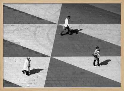A high-angle, black and white photograph capturing three people walking across a paved plaza, their paths intersecting with a dramatic geometric pattern of light and shadow. The strong sunlight creates large, sharp-edged triangles of shade, isolating each figure in the composition, which is enclosed in a silver frame. Decor
