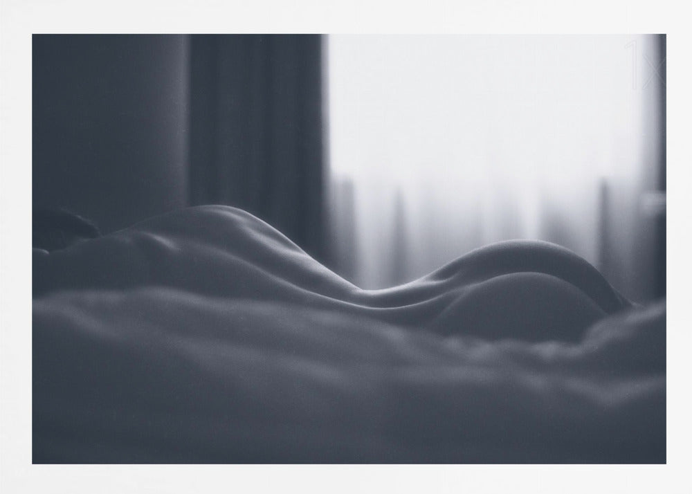 A close-up black and white artistic photograph capturing the soft curves of a person's back and hips as they lie down. The background is softly lit by a window, creating a play of light and shadow that emphasizes the human form's abstract, sculptural qualities. The image is displayed in a silver frame. Print