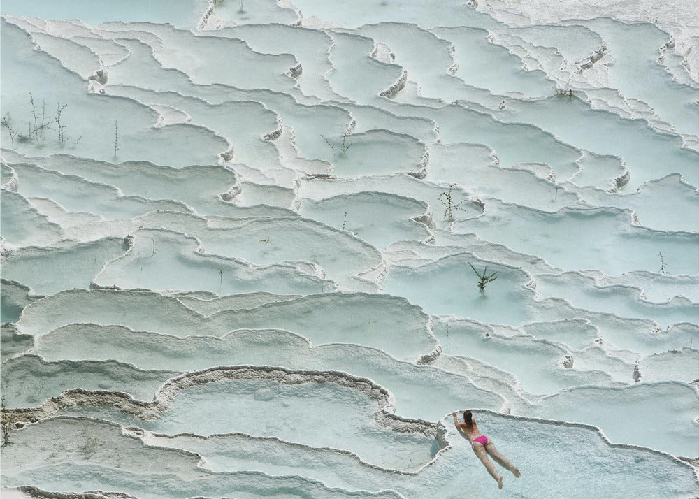 An aerial view of a woman in a pink bikini swimming in the stunning, terraced, milky-blue thermal pools of Pamukkale, Turkey. The white travertine formations create a unique, layered landscape, and the entire image is enclosed in a silver frame. Artwork
