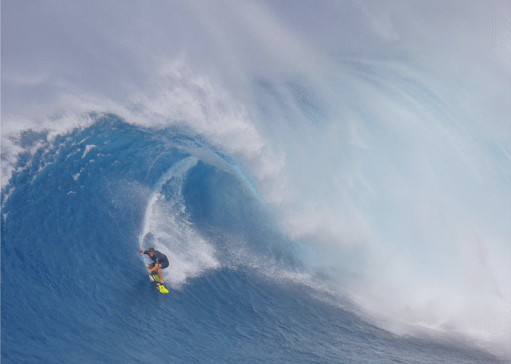 A framed photograph of a professional surfer skillfully riding down the steep face of a massive, cresting blue ocean wave, surrounded by white sea spray. Decor