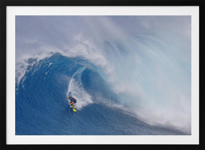 A framed photograph of a professional surfer skillfully riding down the steep face of a massive, cresting blue ocean wave, surrounded by white sea spray. Decor