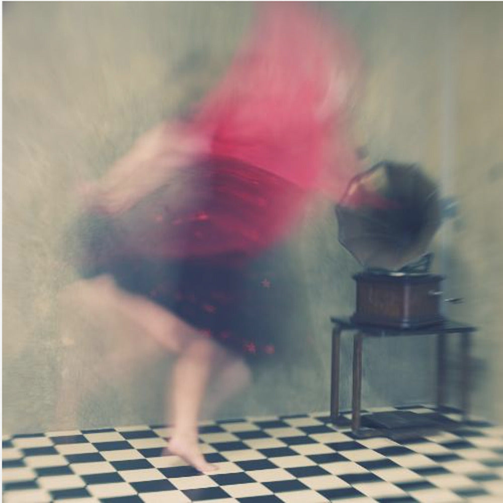 A motion-blurred, artistic photograph in a light wood frame showing a woman dancing barefoot on a black and white checkered floor. She wears a bright red top and a dark skirt, both blurred by her movement. Next to her stands a vintage gramophone on a small table, against a muted, textured wall, creating a dreamy, nostalgic atmosphere. Print