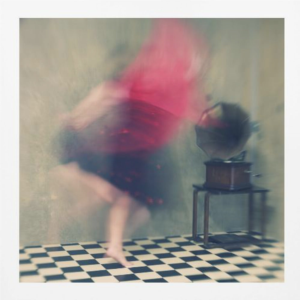 A motion-blurred, artistic photograph in a light wood frame showing a woman dancing barefoot on a black and white checkered floor. She wears a bright red top and a dark skirt, both blurred by her movement. Next to her stands a vintage gramophone on a small table, against a muted, textured wall, creating a dreamy, nostalgic atmosphere. Print