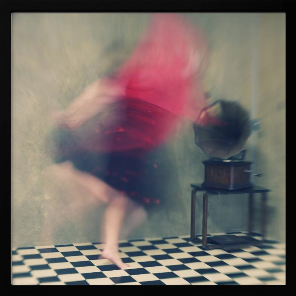 A motion-blurred, artistic photograph in a light wood frame showing a woman dancing barefoot on a black and white checkered floor. She wears a bright red top and a dark skirt, both blurred by her movement. Next to her stands a vintage gramophone on a small table, against a muted, textured wall, creating a dreamy, nostalgic atmosphere. Print