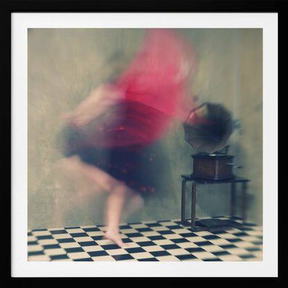 A motion-blurred, artistic photograph in a light wood frame showing a woman dancing barefoot on a black and white checkered floor. She wears a bright red top and a dark skirt, both blurred by her movement. Next to her stands a vintage gramophone on a small table, against a muted, textured wall, creating a dreamy, nostalgic atmosphere. Print