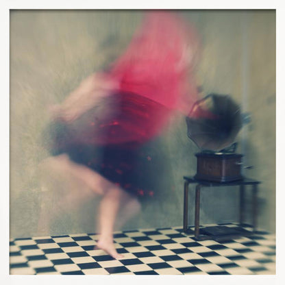 A motion-blurred, artistic photograph in a light wood frame showing a woman dancing barefoot on a black and white checkered floor. She wears a bright red top and a dark skirt, both blurred by her movement. Next to her stands a vintage gramophone on a small table, against a muted, textured wall, creating a dreamy, nostalgic atmosphere. Print