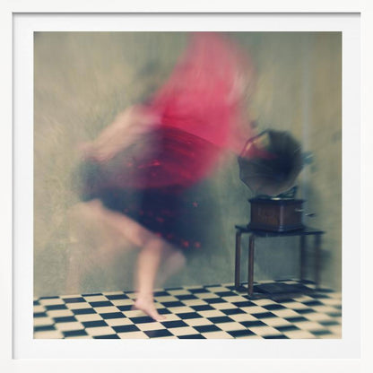 A motion-blurred, artistic photograph in a light wood frame showing a woman dancing barefoot on a black and white checkered floor. She wears a bright red top and a dark skirt, both blurred by her movement. Next to her stands a vintage gramophone on a small table, against a muted, textured wall, creating a dreamy, nostalgic atmosphere. Print