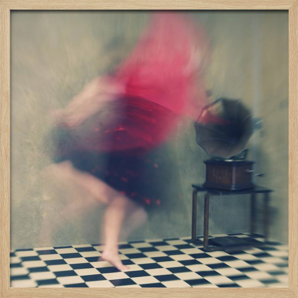 A motion-blurred, artistic photograph in a light wood frame showing a woman dancing barefoot on a black and white checkered floor. She wears a bright red top and a dark skirt, both blurred by her movement. Next to her stands a vintage gramophone on a small table, against a muted, textured wall, creating a dreamy, nostalgic atmosphere. Print