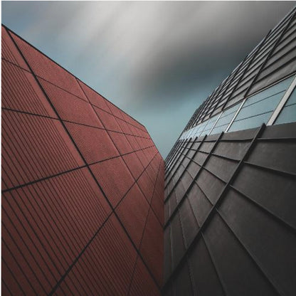 A dramatic low-angle photograph of two modern buildings with contrasting facades. On the left, a building is covered in deep red square panels. On the right, a darker building features black angular tiles and glass windows. The sky above is a moody, blurred grey. Print