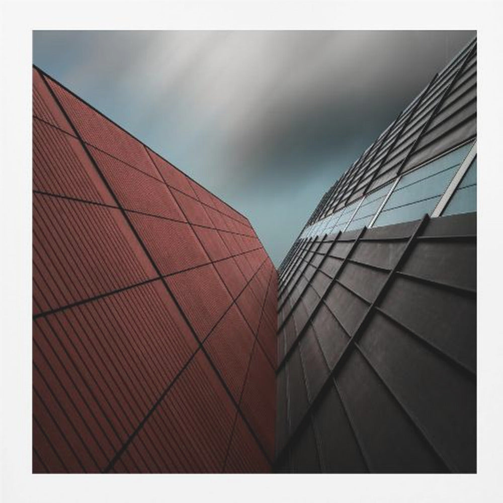 A dramatic low-angle photograph of two modern buildings with contrasting facades. On the left, a building is covered in deep red square panels. On the right, a darker building features black angular tiles and glass windows. The sky above is a moody, blurred grey. Print