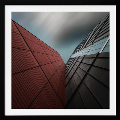 A dramatic low-angle photograph of two modern buildings with contrasting facades. On the left, a building is covered in deep red square panels. On the right, a darker building features black angular tiles and glass windows. The sky above is a moody, blurred grey. Print