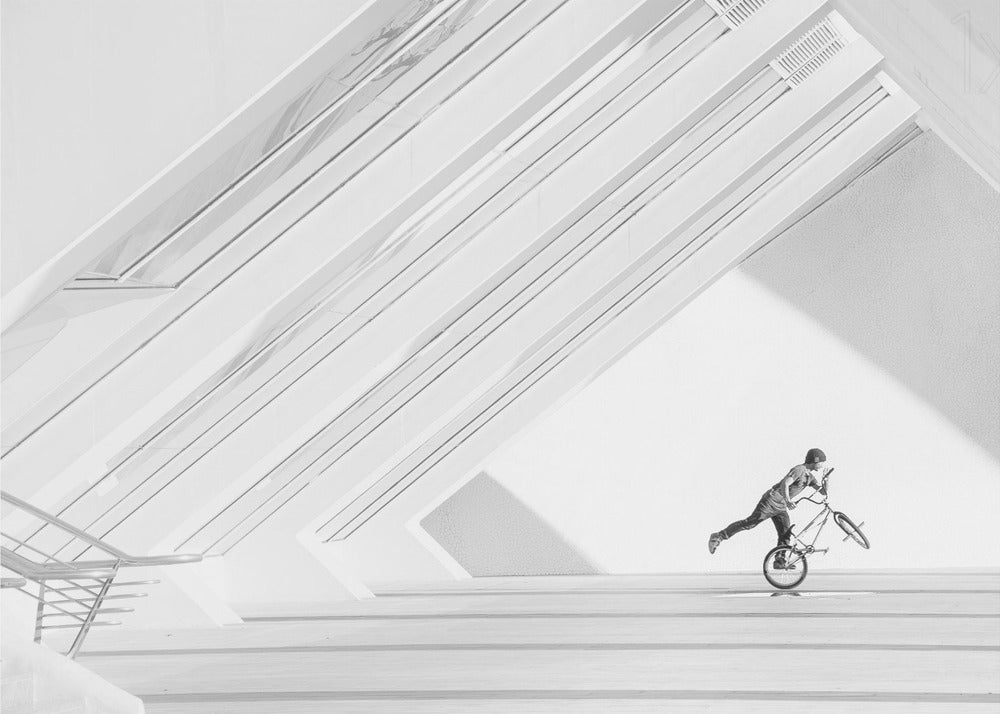 A minimalist black and white photograph in a silver frame, featuring a lone BMX rider performing a trick on the back wheel inside a stark, modern building with strong diagonal architectural lines. Poster