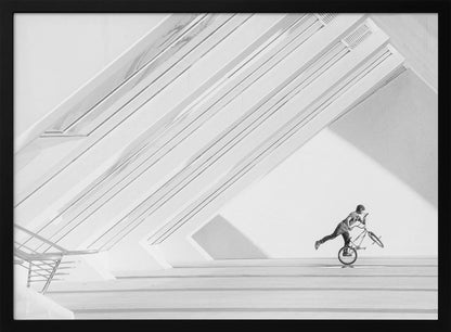 A minimalist black and white photograph in a silver frame, featuring a lone BMX rider performing a trick on the back wheel inside a stark, modern building with strong diagonal architectural lines. Poster