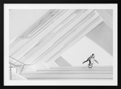 A minimalist black and white photograph in a silver frame, featuring a lone BMX rider performing a trick on the back wheel inside a stark, modern building with strong diagonal architectural lines. Poster