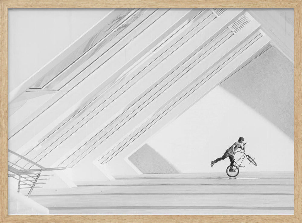 A minimalist black and white photograph in a silver frame, featuring a lone BMX rider performing a trick on the back wheel inside a stark, modern building with strong diagonal architectural lines. Poster