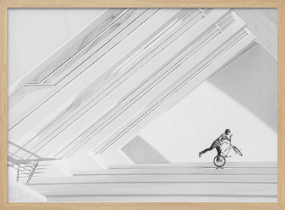 A minimalist black and white photograph in a silver frame, featuring a lone BMX rider performing a trick on the back wheel inside a stark, modern building with strong diagonal architectural lines. Poster