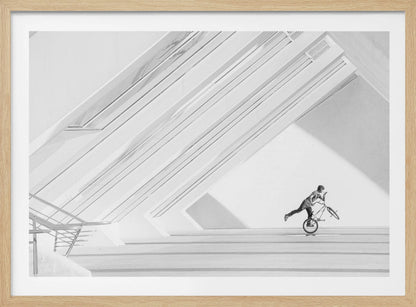 A minimalist black and white photograph in a silver frame, featuring a lone BMX rider performing a trick on the back wheel inside a stark, modern building with strong diagonal architectural lines. Poster