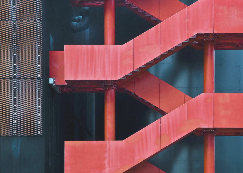 A framed abstract architectural photograph of a bright red industrial staircase zig-zagging against a dark teal wall. The composition highlights the geometric shapes and bold color contrast between the red stairs and the cool-toned background, with a silver mesh panel visible on the left side. Print