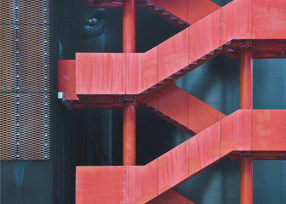 A framed abstract architectural photograph of a bright red industrial staircase zig-zagging against a dark teal wall. The composition highlights the geometric shapes and bold color contrast between the red stairs and the cool-toned background, with a silver mesh panel visible on the left side. Print