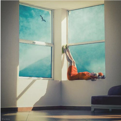 A surreal photograph of a woman in a bright orange dress lying on a wide, sunlit windowsill with her legs up against the wall. Through the large windows, a single bird is seen flying in a vibrant teal sky. The image, presented in a light wood frame, captures a moment of quiet contemplation and escape. Wall Art