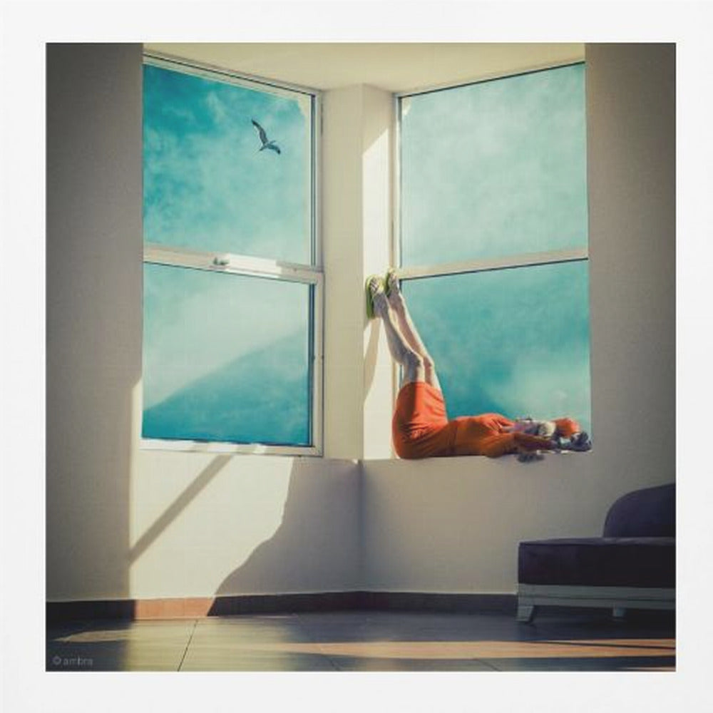 A surreal photograph of a woman in a bright orange dress lying on a wide, sunlit windowsill with her legs up against the wall. Through the large windows, a single bird is seen flying in a vibrant teal sky. The image, presented in a light wood frame, captures a moment of quiet contemplation and escape. Wall Art