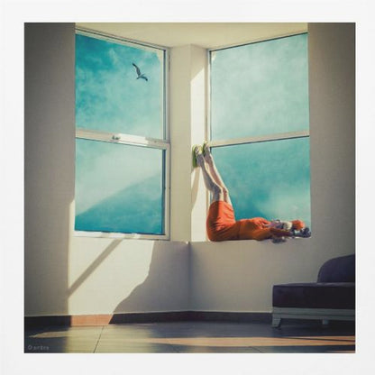 A surreal photograph of a woman in a bright orange dress lying on a wide, sunlit windowsill with her legs up against the wall. Through the large windows, a single bird is seen flying in a vibrant teal sky. The image, presented in a light wood frame, captures a moment of quiet contemplation and escape. Wall Art
