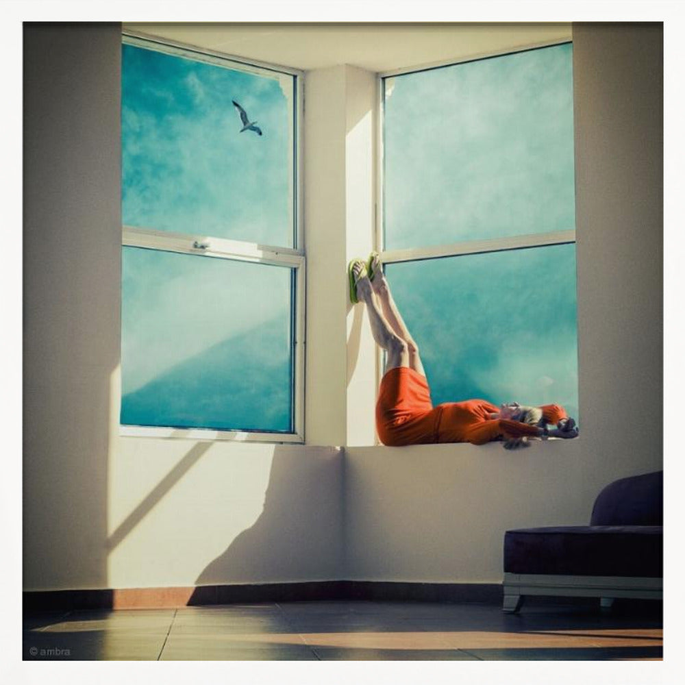 A surreal photograph of a woman in a bright orange dress lying on a wide, sunlit windowsill with her legs up against the wall. Through the large windows, a single bird is seen flying in a vibrant teal sky. The image, presented in a light wood frame, captures a moment of quiet contemplation and escape. Wall Art