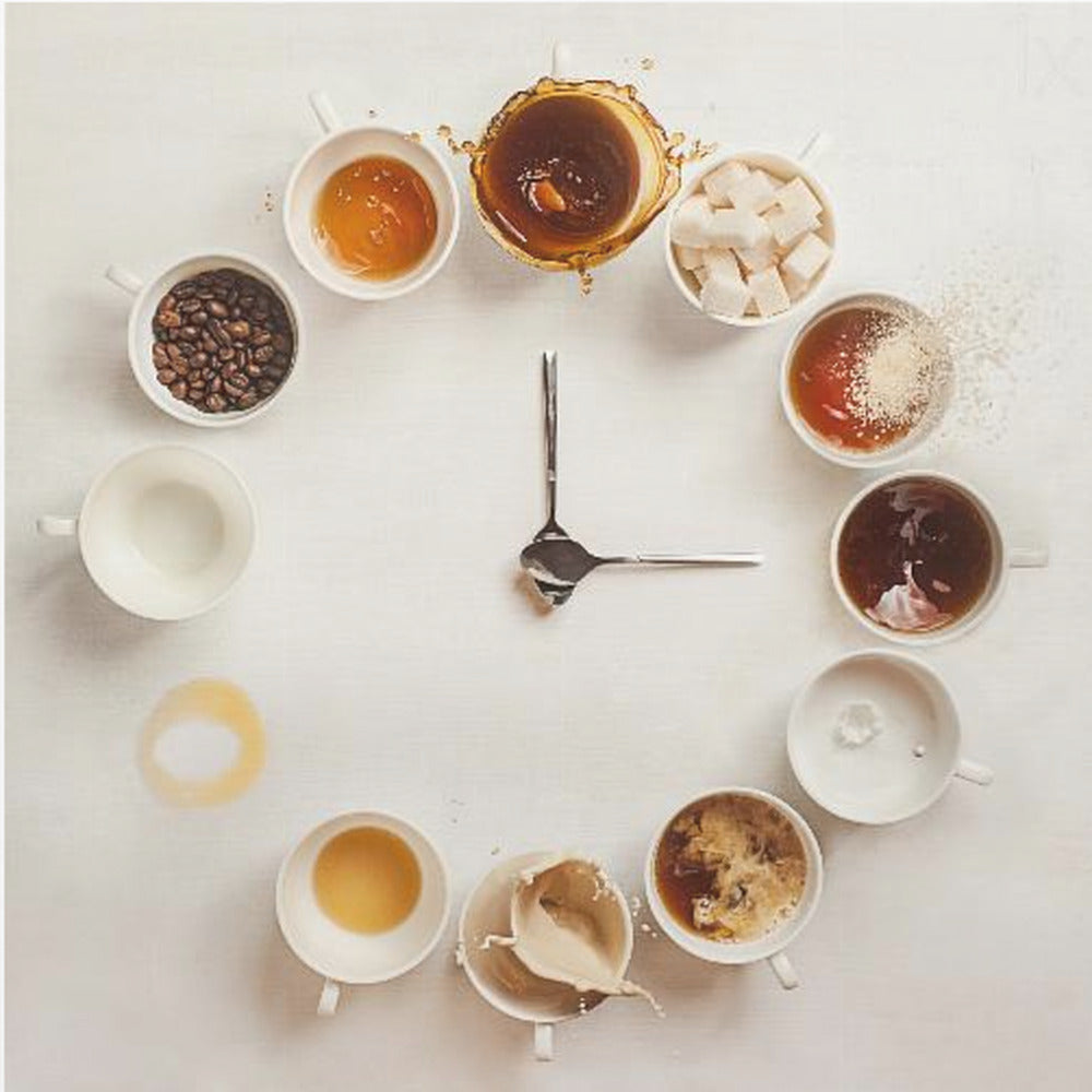 A creative flat lay photograph of a clock made from coffee cups and ingredients. Twelve white cups are arranged in a circle on a white background, each representing an hour and filled with different coffee-related items like beans, honey, sugar cubes, and various stages of coffee with milk splashing. In the center, two silver spoons form the hands of the clock. The entire image is presented within a light wood frame. Decor