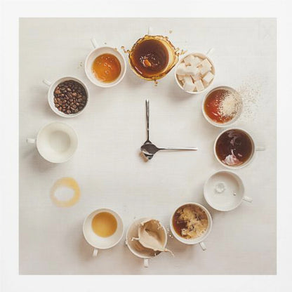 A creative flat lay photograph of a clock made from coffee cups and ingredients. Twelve white cups are arranged in a circle on a white background, each representing an hour and filled with different coffee-related items like beans, honey, sugar cubes, and various stages of coffee with milk splashing. In the center, two silver spoons form the hands of the clock. The entire image is presented within a light wood frame. Decor