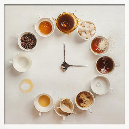 A creative flat lay photograph of a clock made from coffee cups and ingredients. Twelve white cups are arranged in a circle on a white background, each representing an hour and filled with different coffee-related items like beans, honey, sugar cubes, and various stages of coffee with milk splashing. In the center, two silver spoons form the hands of the clock. The entire image is presented within a light wood frame. Decor