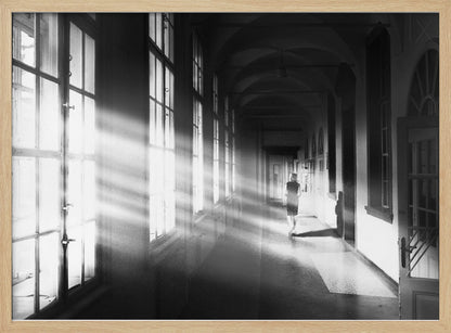 A black and white photograph of a woman walking away down a long, dark hallway. Bright shafts of sunlight pour through large windows on the left, creating a dramatic interplay of light and shadow on the floor and walls. Poster