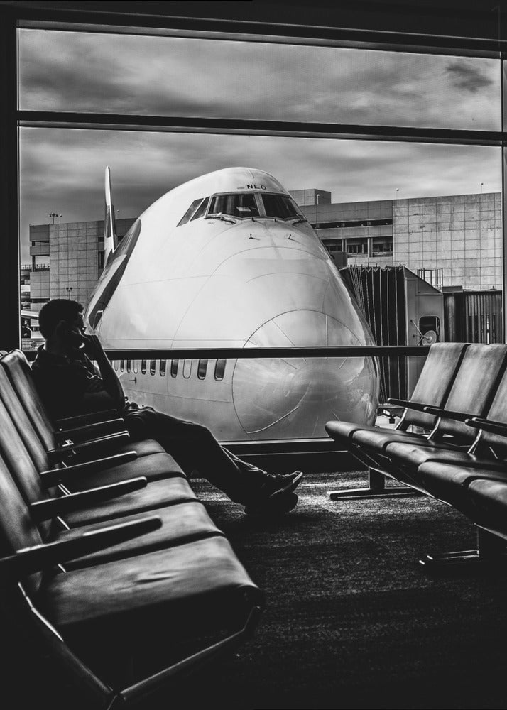 A dramatic black and white photograph of a silhouetted person sitting in an airport departure lounge, waiting for their flight. Through the large window, the enormous nose of a Boeing 747 jumbo jet is visible up close, dominating the view under a cloudy sky. Artwork