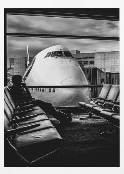 A dramatic black and white photograph of a silhouetted person sitting in an airport departure lounge, waiting for their flight. Through the large window, the enormous nose of a Boeing 747 jumbo jet is visible up close, dominating the view under a cloudy sky. Artwork