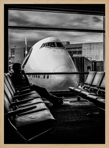 A dramatic black and white photograph of a silhouetted person sitting in an airport departure lounge, waiting for their flight. Through the large window, the enormous nose of a Boeing 747 jumbo jet is visible up close, dominating the view under a cloudy sky. Artwork