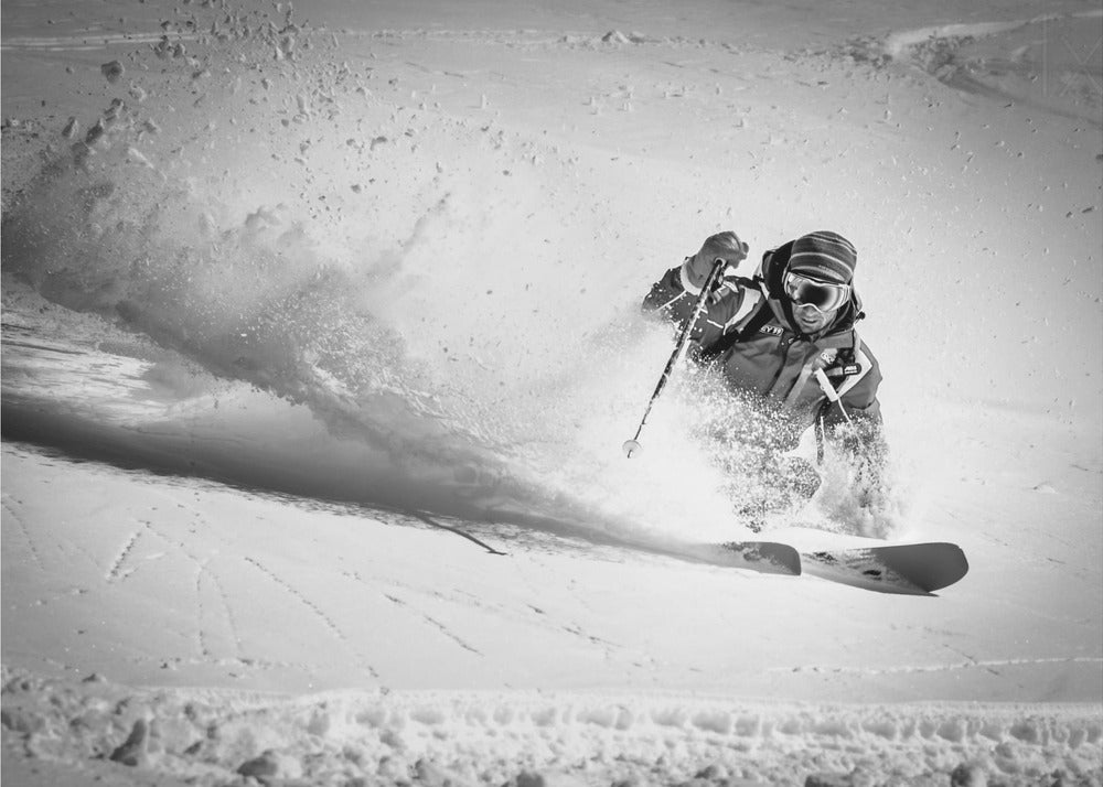 A framed, black and white action shot of a skier making a sharp turn on a snowy slope, sending a large spray of powder into the air. The skier is leaning into the turn, fully kitted out in winter gear including goggles and a beanie. Artwork